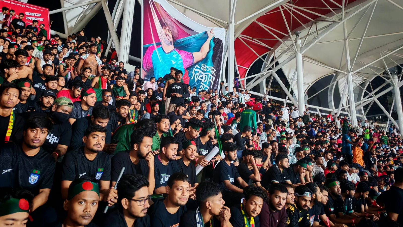 Supporters of the block in black in the stand at night, a player's banner above them.